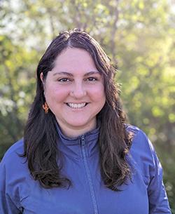 Michelle stands in front of a green-leaved bush, wearing a blue zippered jacket